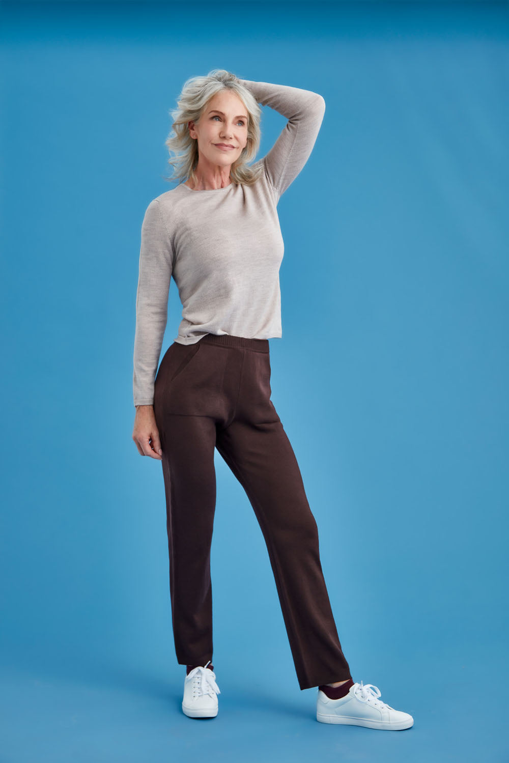 Woman posing in a gray sweater and brown merino pants against a blue background