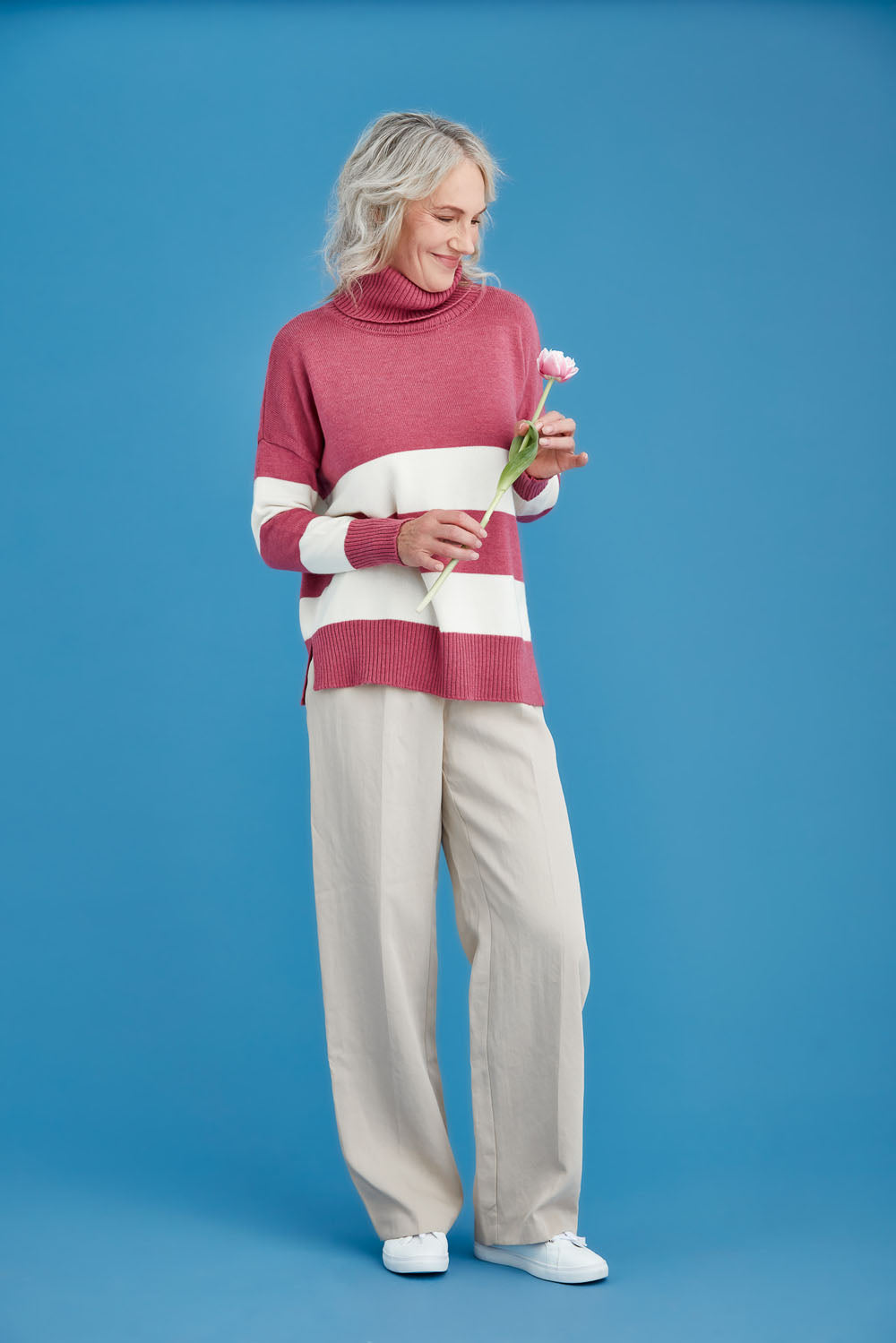 Woman wearing a striped merino sweater and beige pants holding a flower against a blue background