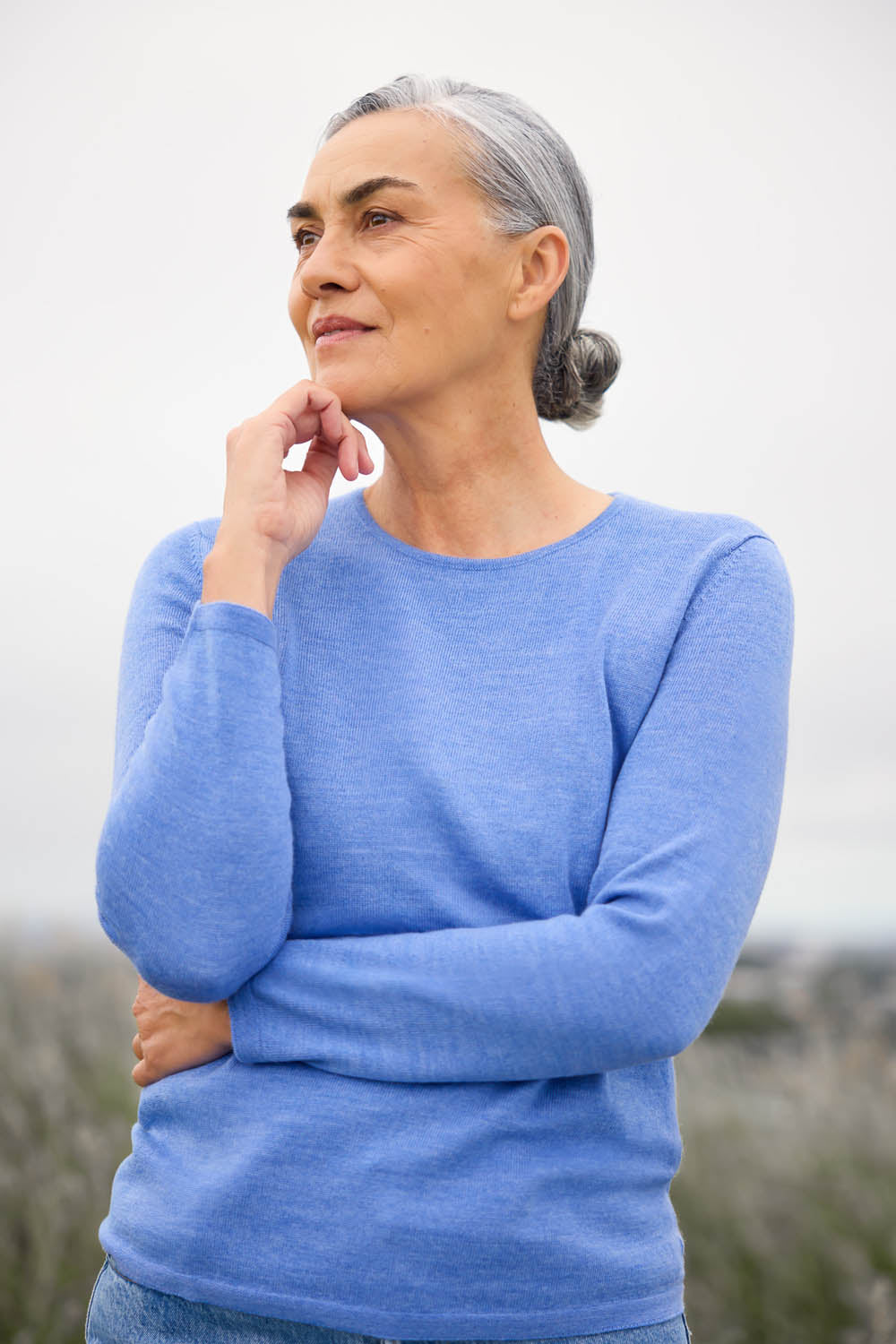 A woman standing in an outdoor setting, wearing a light blue crew neck jumper and blue jeans.