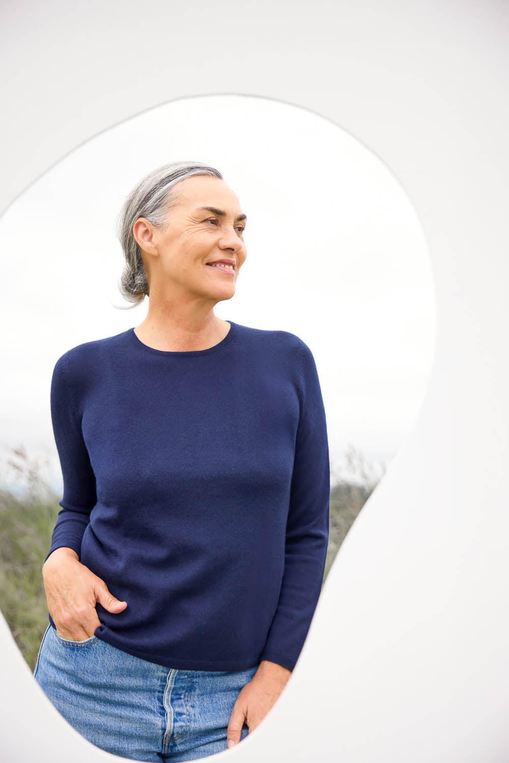 A woman standing in an outdoor setting, wearing a blue crew neck jumper and blue jeans.