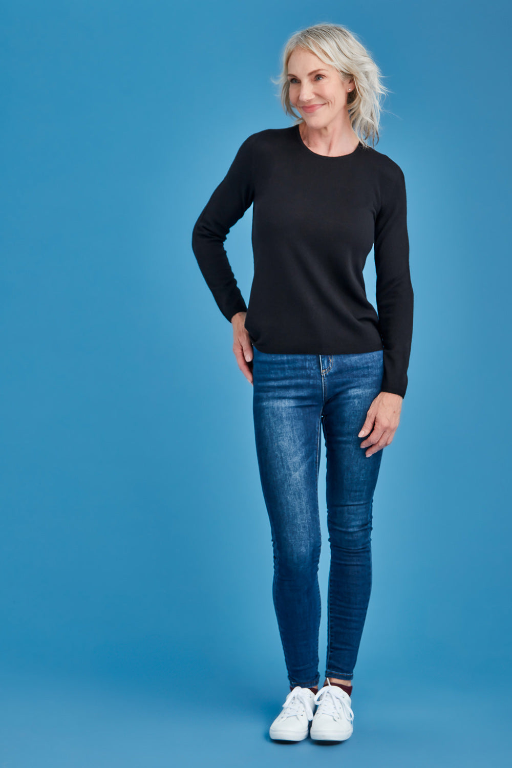 Woman wearing a black long-sleeve merino shirt and blue jeans on a blue background