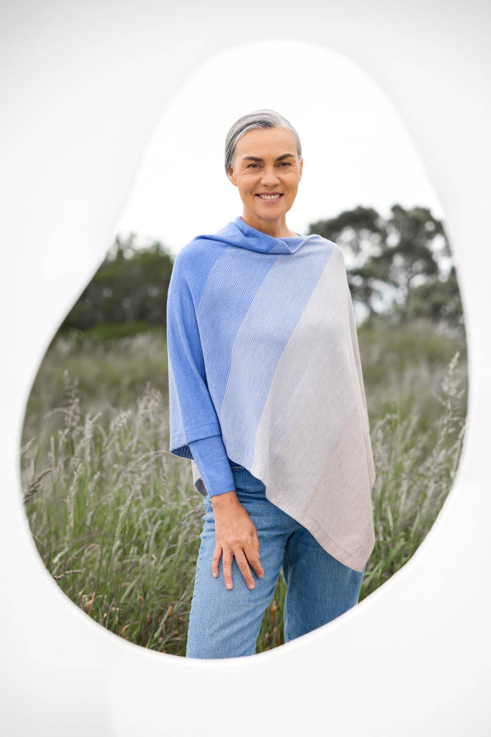 A woman standing in a field with a graduated stripe poncho featuring ombre stripes in blue and beige draped over her shoulders.