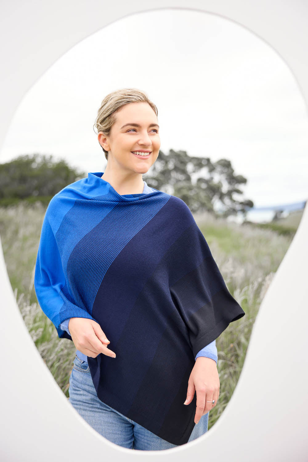 A woman standing in a field with a graduated stripe poncho featuring ombre stripes in blue draped over her shoulders.