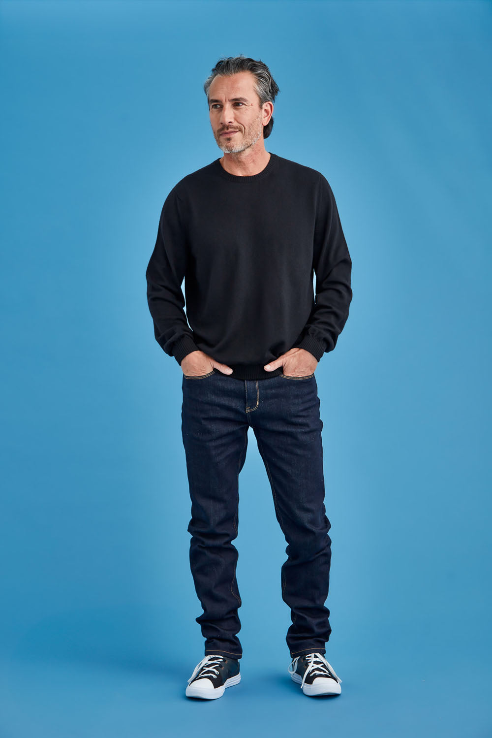 Man wearing a black sweater and jeans standing against a blue background