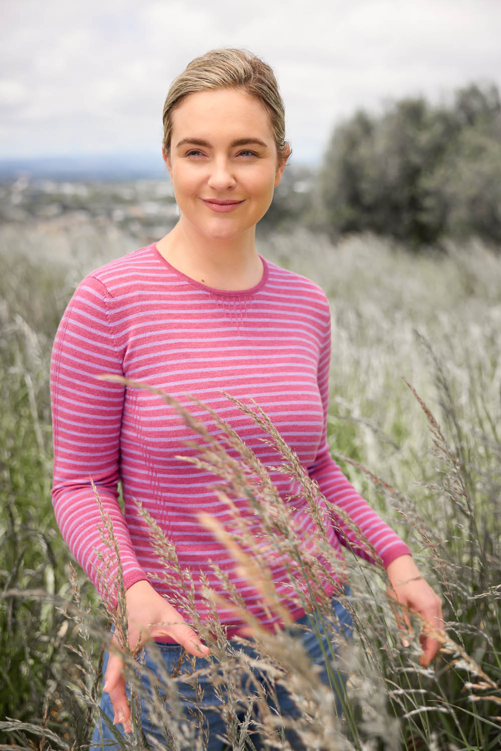 A woman standing in a field of dry grass wearing a pink striped jumper and blue jeans.