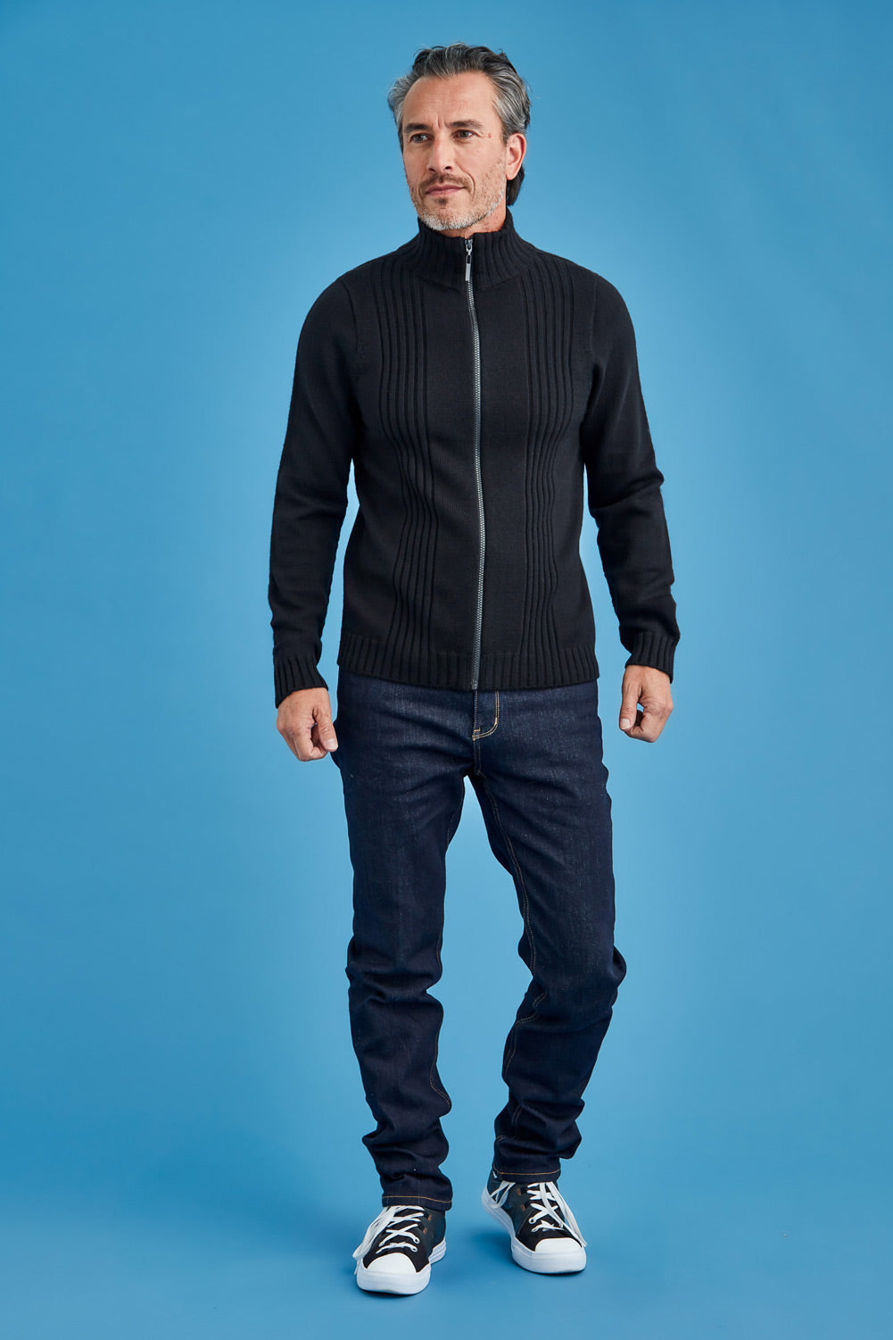 Man wearing a black zip-up sweater and dark jeans against a blue background